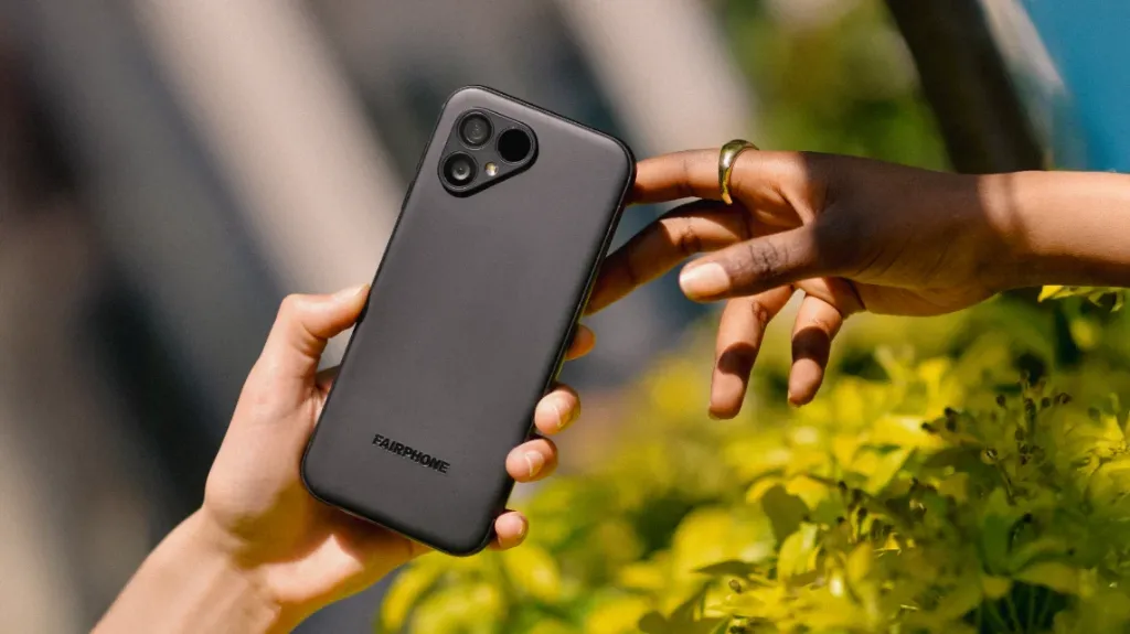 Two hands exchanging a Fairphone 5 with the Fairphone logo on the back, set against a backdrop of leafy plants, emphasising climate-conscious design.